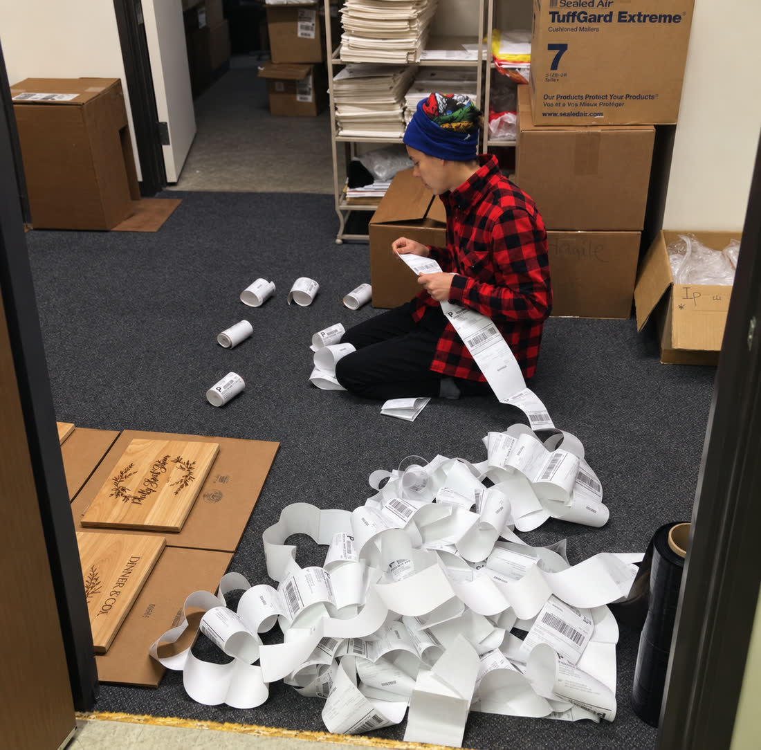 A person kneeling on an office floor, sorting a long, tangled river of printed shipping labels during a busy season